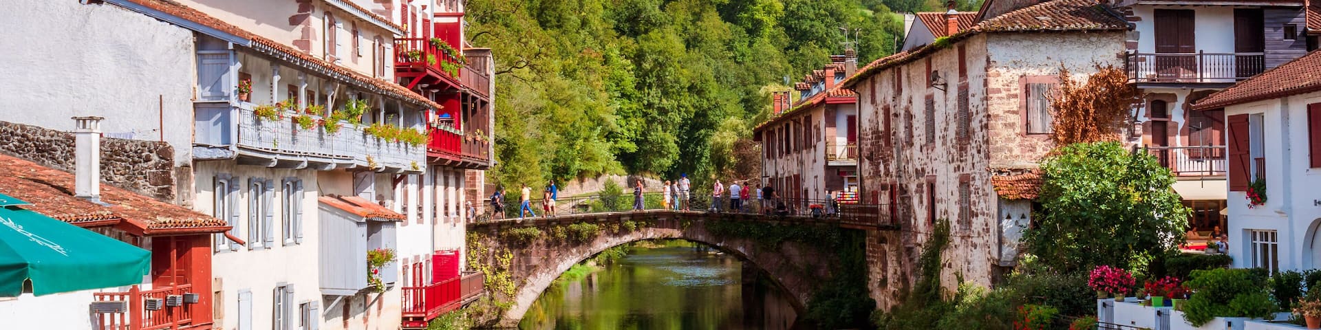 A river with a stone bridge, buildings with balconies, and lush greenery in the background.