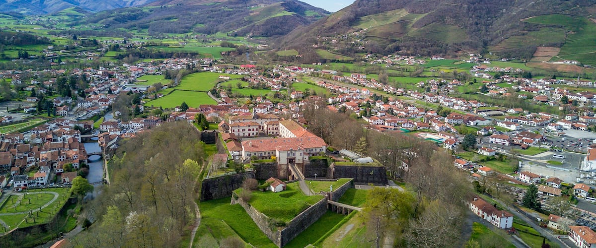 A town nestled in a valley with a prominent castle-like structure surrounded by lush green fields and mountains in the background.