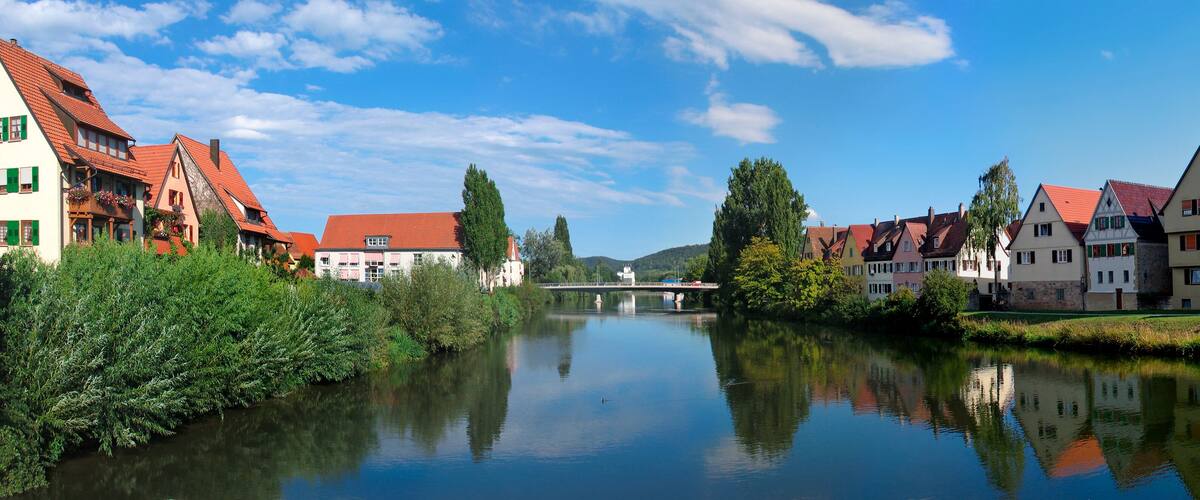A calm river with traditional European houses on both sides, a clear blue sky, and a bridge in the distance.