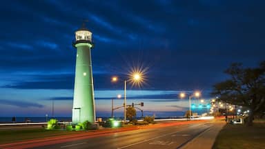 A tall green lighthouse at dusk with street lights and traffic lights in the foreground.