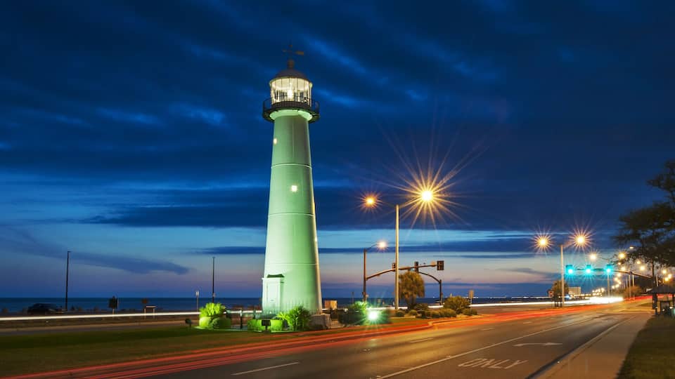 A tall green lighthouse at dusk with street lights and traffic lights in the foreground.