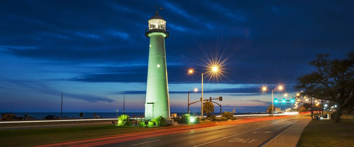 A tall green lighthouse at dusk with street lights and traffic lights in the foreground.