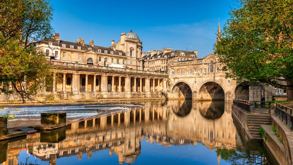 Un bâtiment historique, avec des ponts en arc de cercle enjambant une rivière paisible, dont l’architecture se reflète dans l’eau.