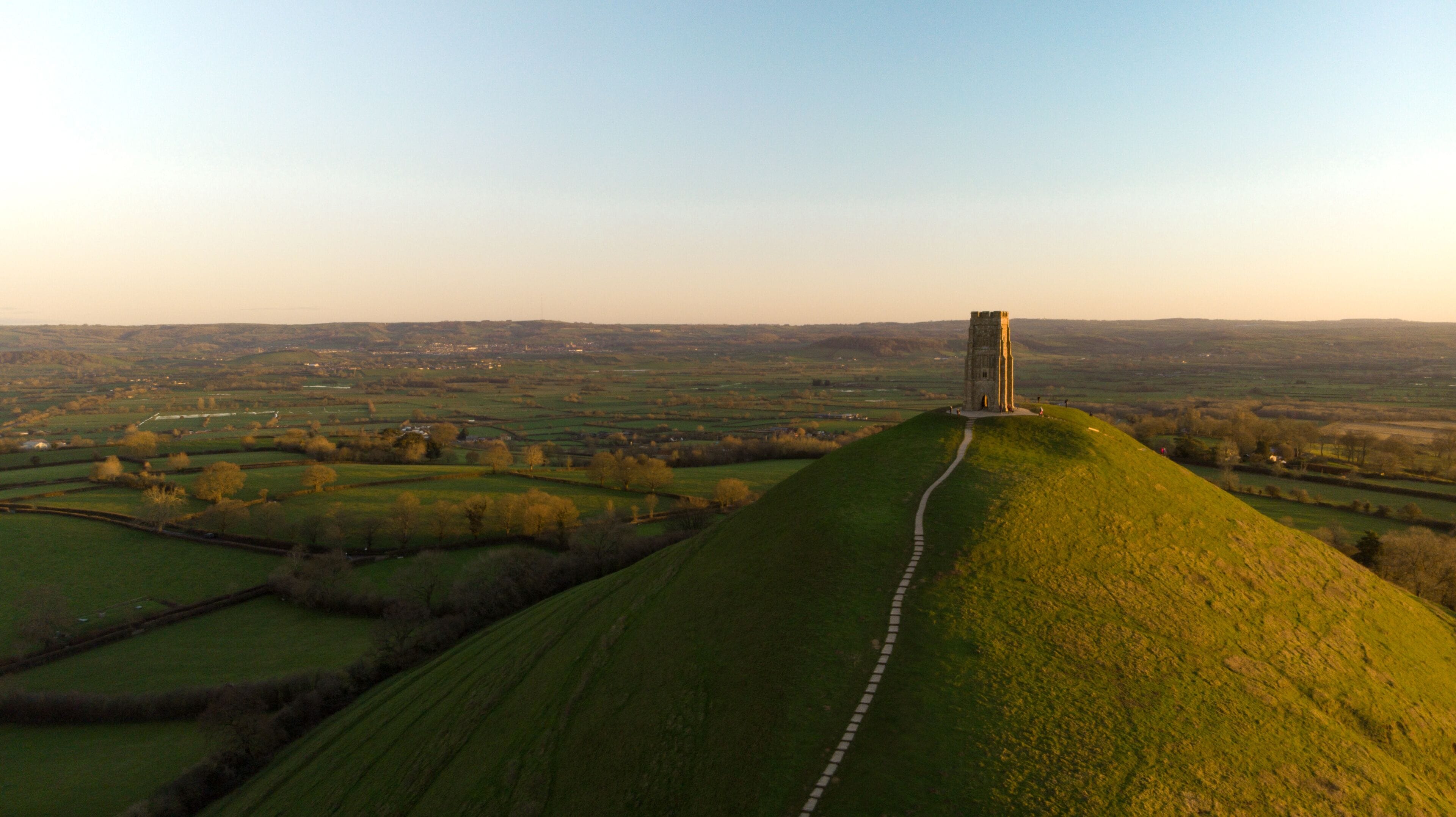 A tower on a grassy hill with a path leading to it, surrounded by fields and distant buildings.