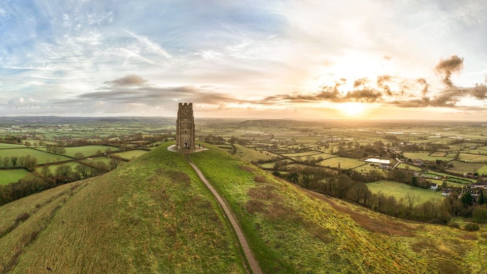 A tower on a hill overlooking a rural landscape with fields and a sunset.