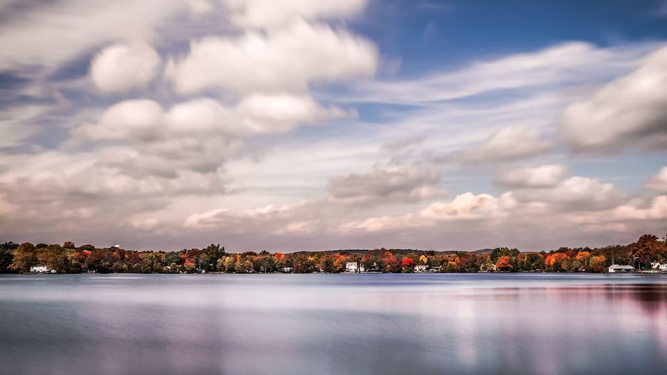 A calm lake with autumn foliage and a cloudy sky.