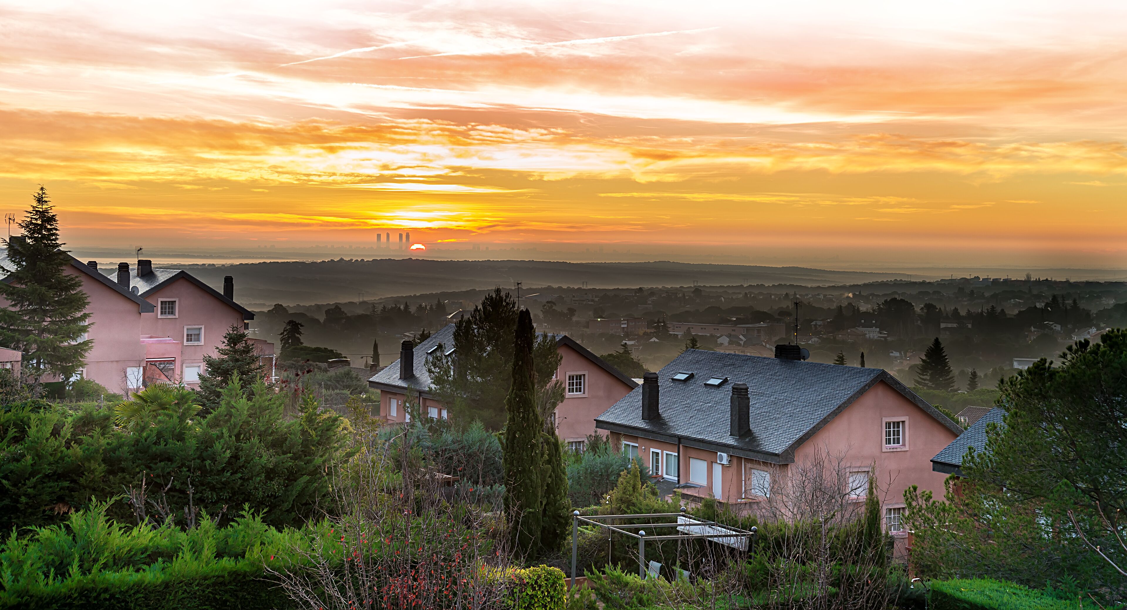 A pink house with a black roof, surrounded by greenery and a sunset in the background.