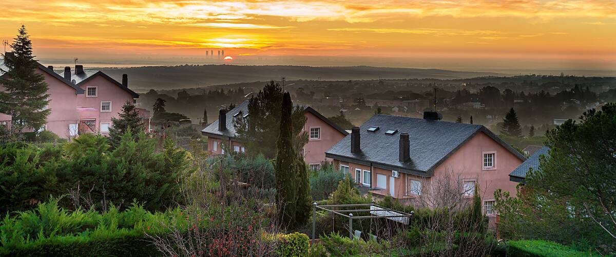 A pink house with a black roof, surrounded by greenery and a sunset in the background.