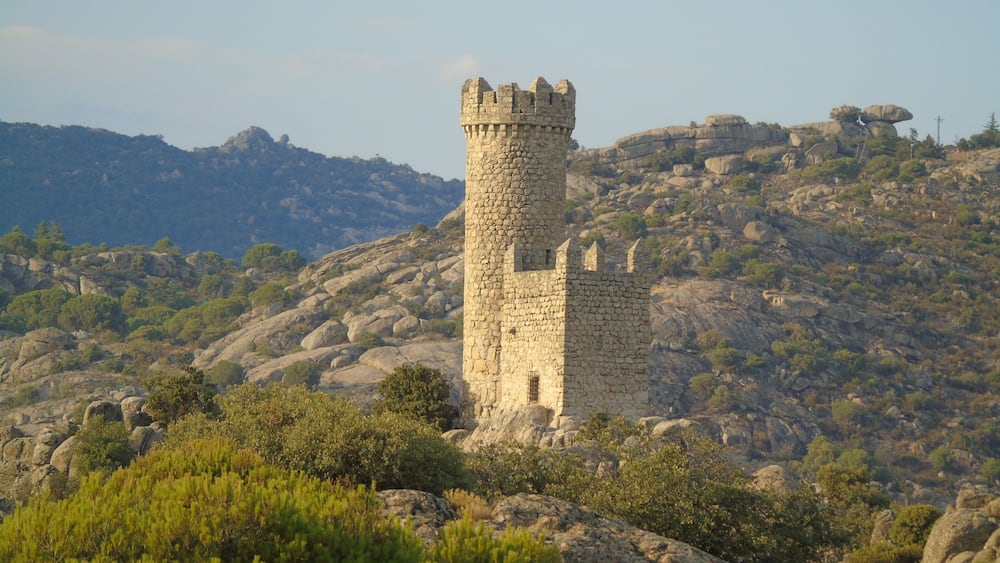 Ein steinerner Turm auf einem felsigen Hügel, umgeben von Vegetation, mit Bergen im Hintergrund.
