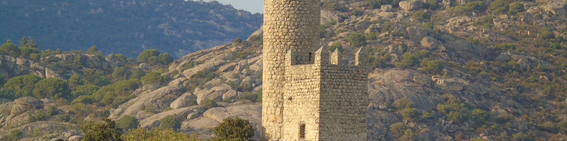 Una torre de piedra sobre una colina rocosa, rodeada de vegetación y con montañas al fondo.