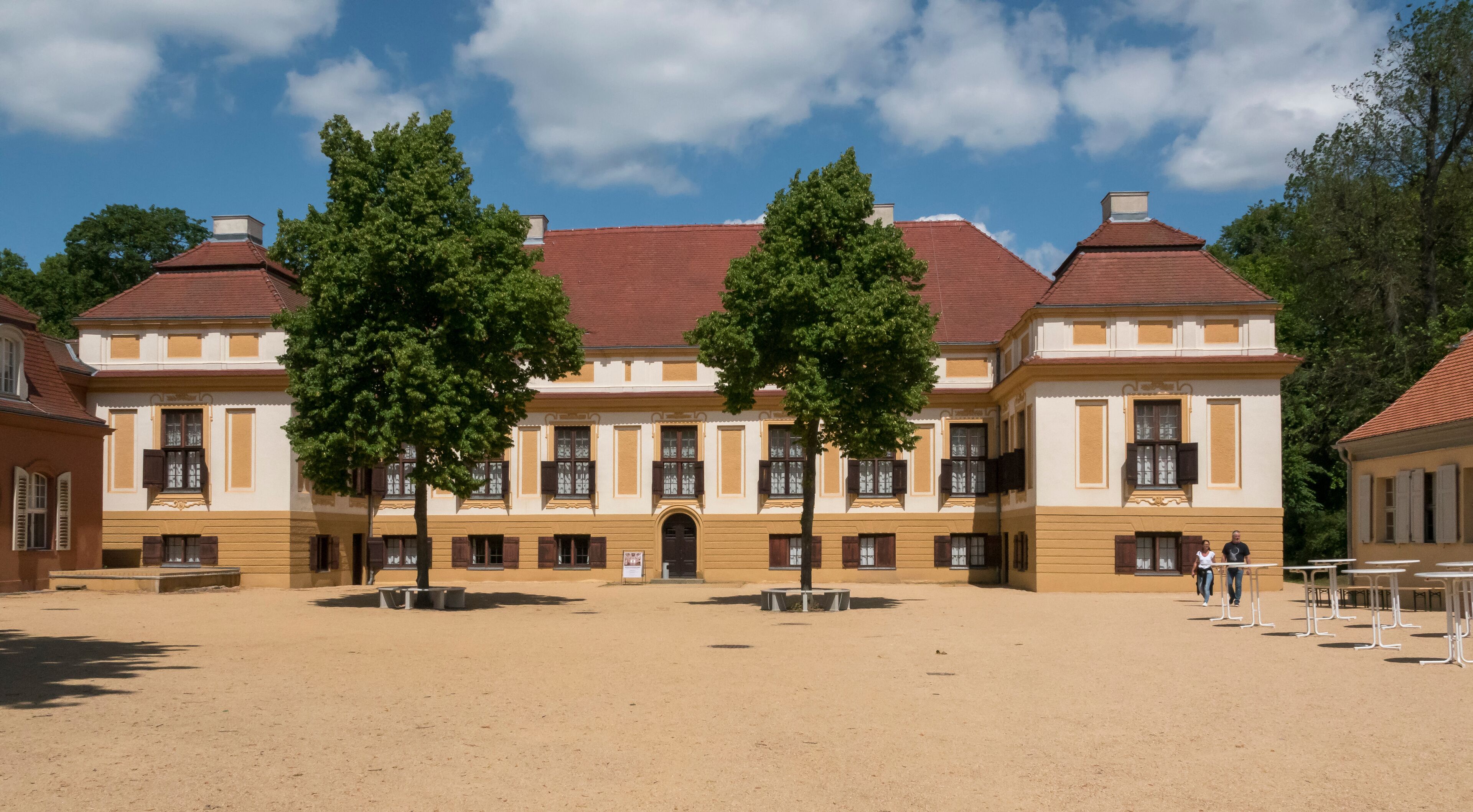 A historic building with a red roof and yellow walls, surrounded by trees and open space.