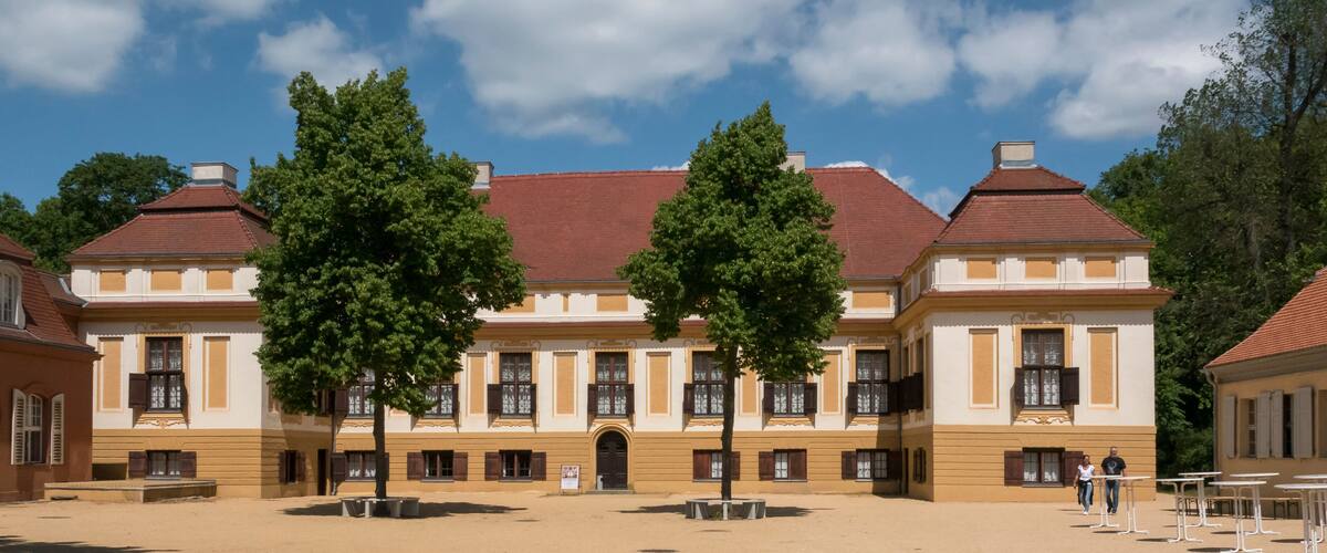 A historic building with a red roof and yellow walls, surrounded by trees and open space.