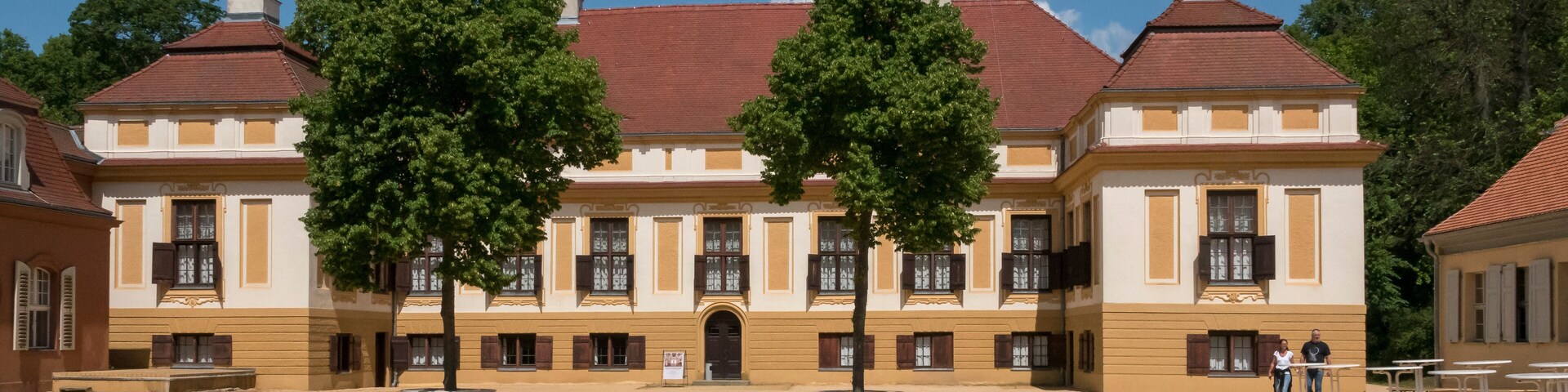 A historic building with a red roof and yellow walls, surrounded by trees and open space.