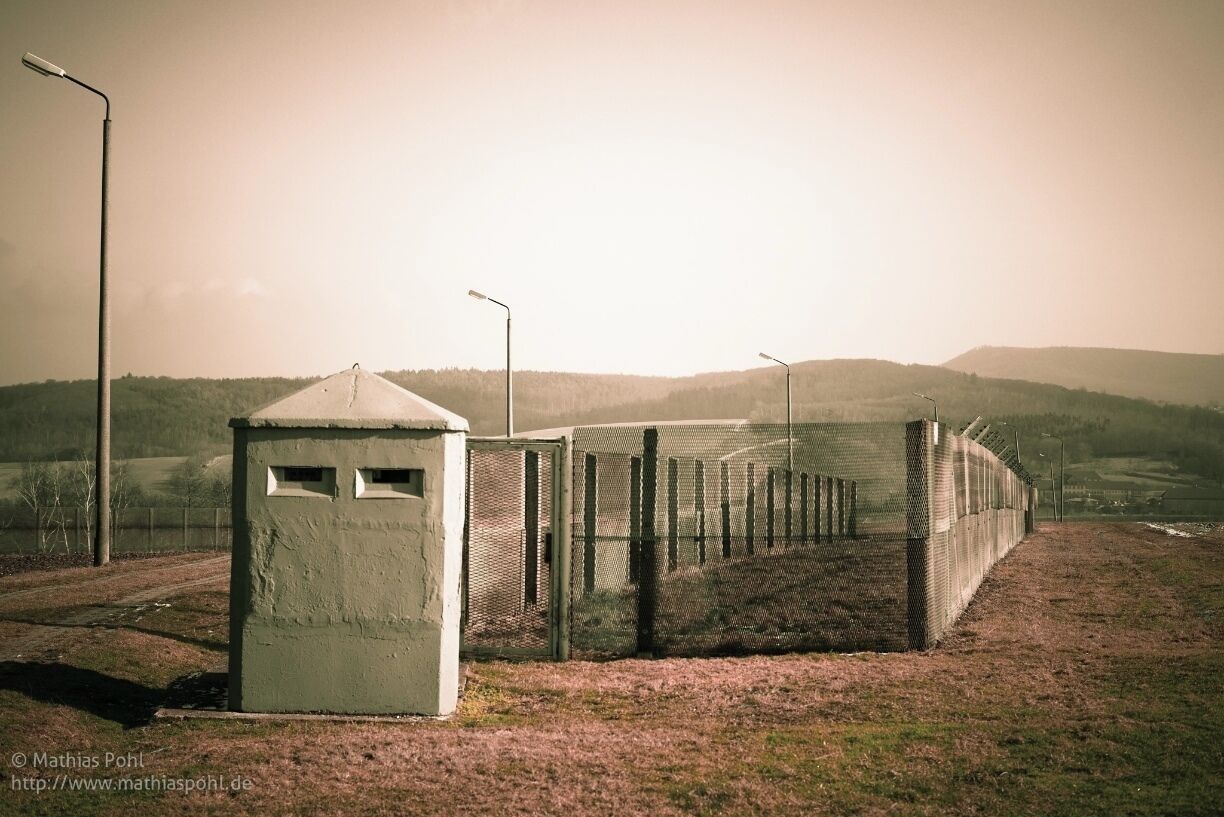 Part #9: Border Dogs and Dog run
The Dog run shown is here a reconstruction. Although there was no run like this in this area, it is typical of dog runs on other sections of the east german border. These were built from 1974, mainly at sections of the border security signal fence that were difficult to guard. Inside of the run were a kennel, a food bowl and a water bowl. There were also "running ropes", fixed between two steel masts across the length of 30 - 50 m. A movable lead was attached to this wire rope, so that the dog could run along the strip. There were gaps between the ends of the ropes to stop the dogs from biting each other.
The guards dogs, which usually spent their entire lives on the border, were fed and checked once a day (taken out of the run every week).