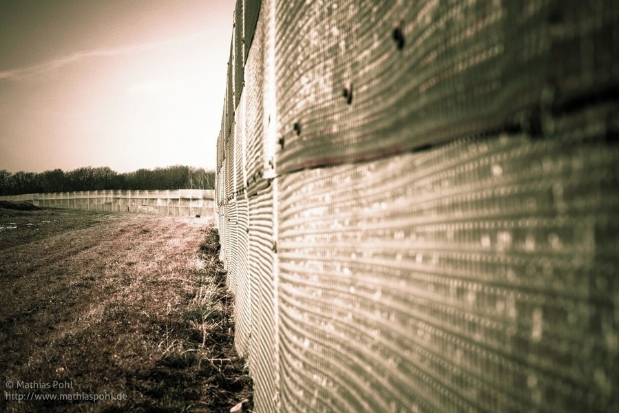 Part #7: Outer Fence
The outer fences were constructed in a number of phases, starting with the initial fortification of the border from May 1952.
This Photo is showing a "third-generation" fence, much more solidly constructed (1960s to the 1980s). The fence line was moved back to create an outer strip between the fence and the actual border. The barbed-wire fences were replaced with a barrier that was usually 3.2â4.0 metres (10â13 ft) high, like this.
The openings in the mesh were generally too small to provide finger-holds and were very sharp. The panels could not easily be pulled down, as they overlapped, and they could not be cut through with a bolt- or wire-cutter. Nor could they be tunnelled under easily, as the bottom segment of the fences was partially buried in the ground.