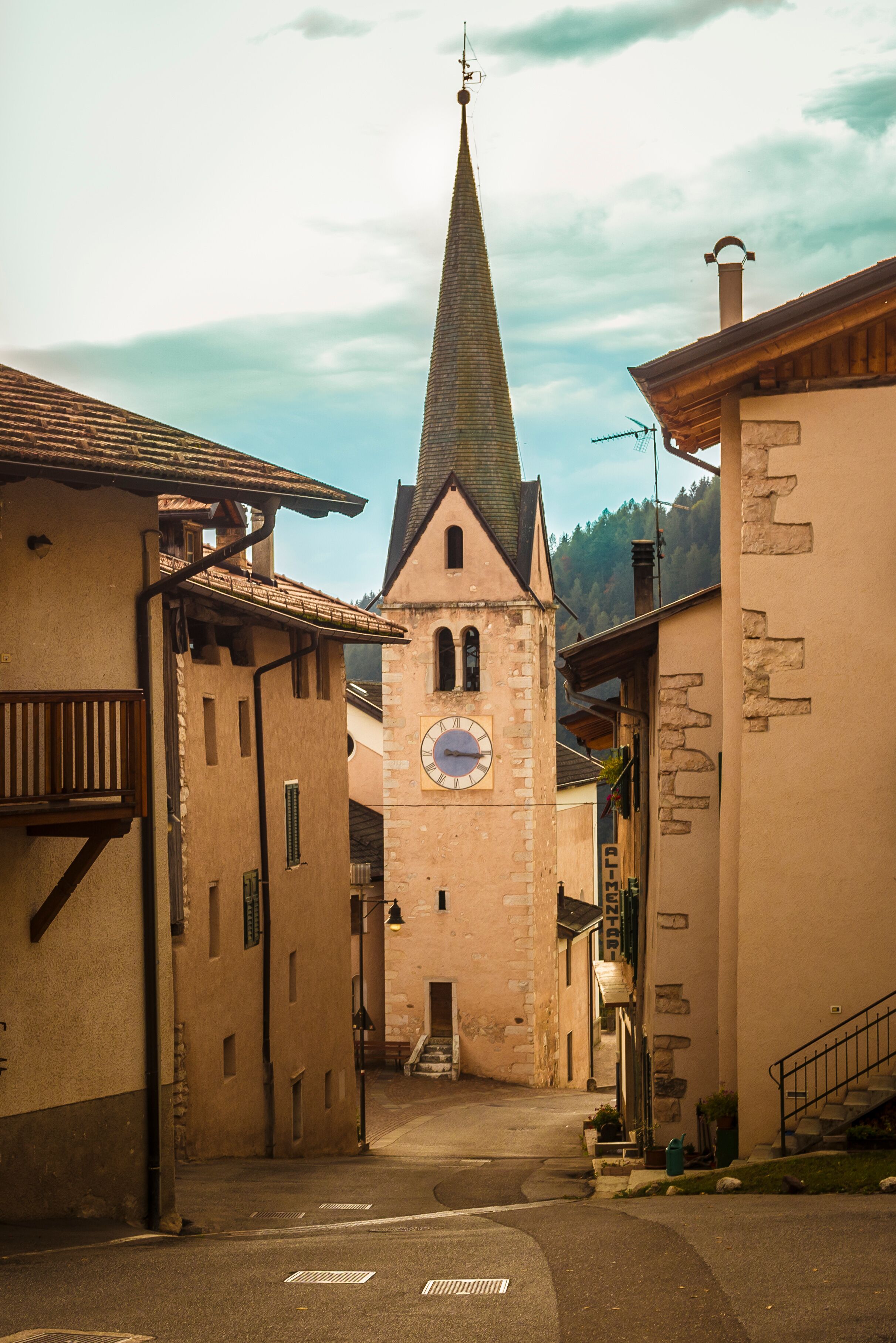 A narrow street with buildings on either side, a church with a clock tower, and a mountain in the background.