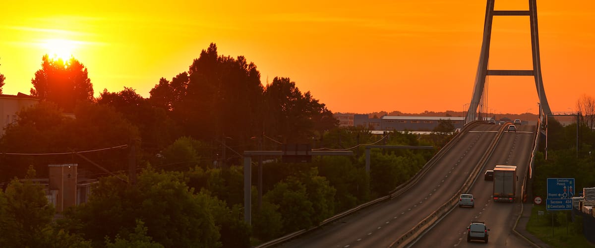 A bridge with a sunset in the background, surrounded by greenery and a highway with vehicles.