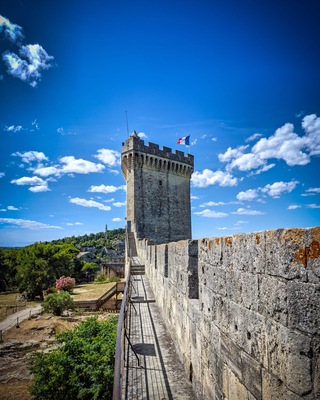 Une tour de pierre historique, surmontée d’un drapeau, un mur de pierre et un ciel bleu dégagé parsemé de nuages.