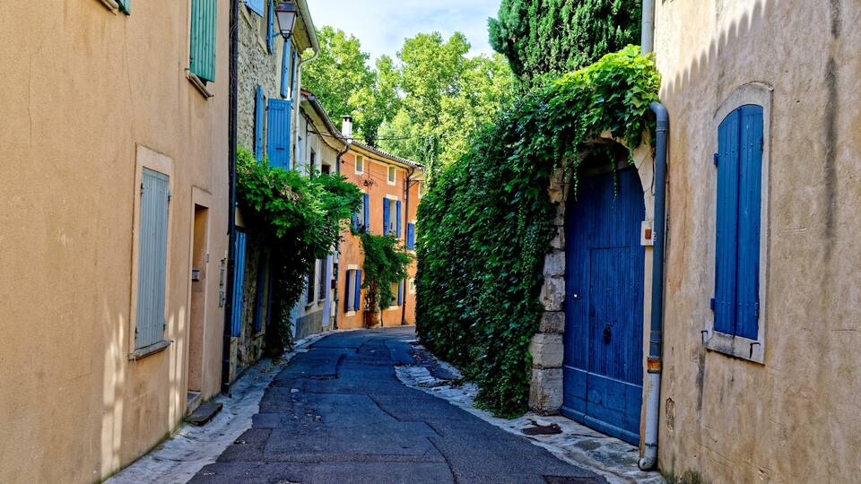 A narrow street flanked by buildings with blue shutters and a tree-lined path.