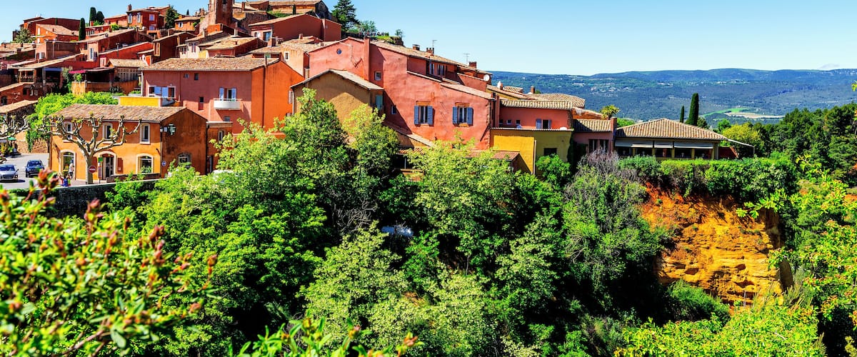 A hillside village with terracotta-roofed buildings, lush greenery, and a clear blue sky.
