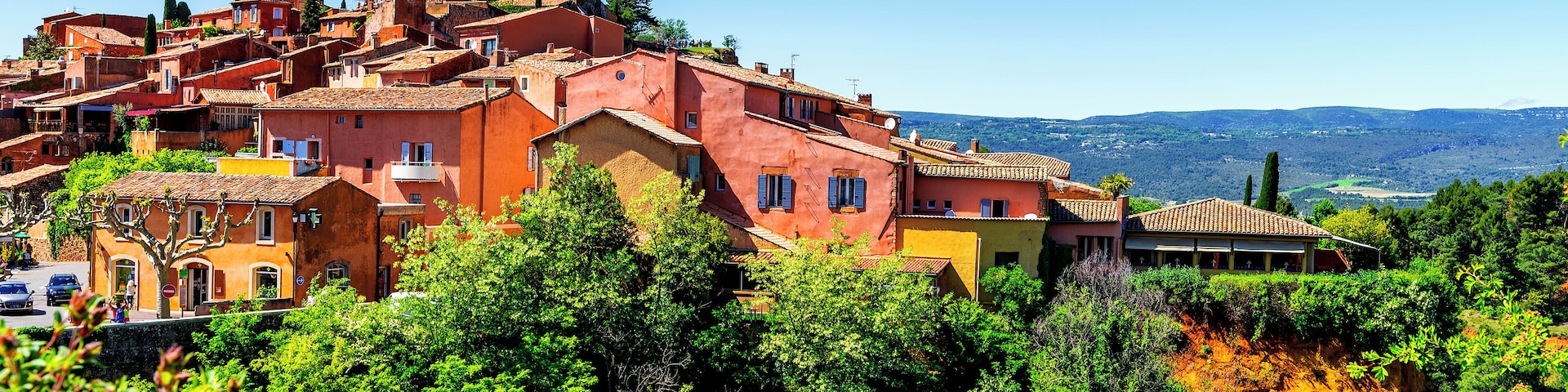 A hillside village with terracotta-roofed buildings, lush greenery, and a clear blue sky.
