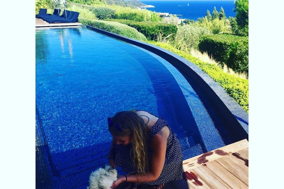Une femme caresse un chien au bord d’une piscine, avec vue sur l’océan et les collines.