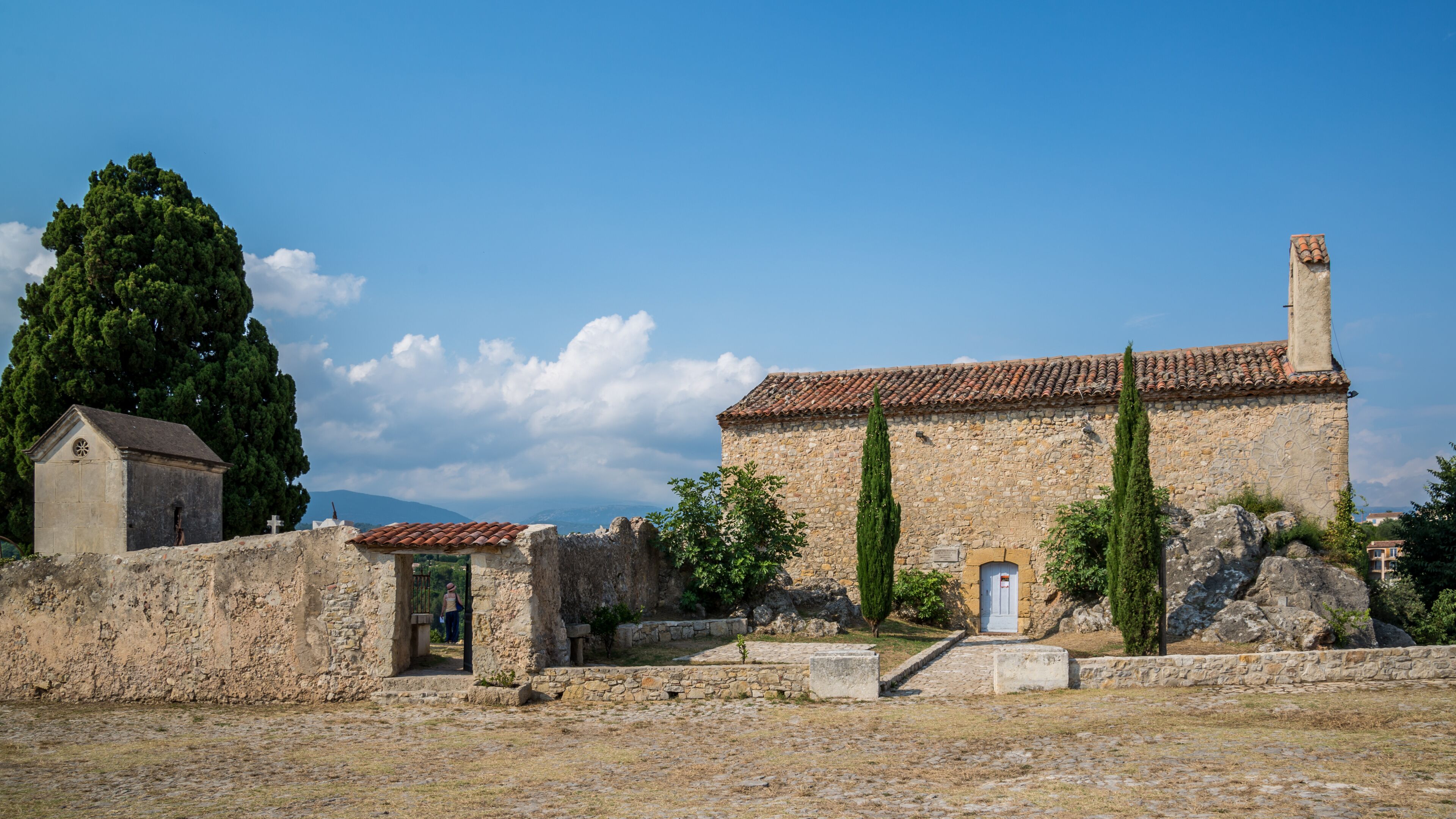 An ancient stone building with a tiled roof, a small church, and a large tree in a rural setting.
