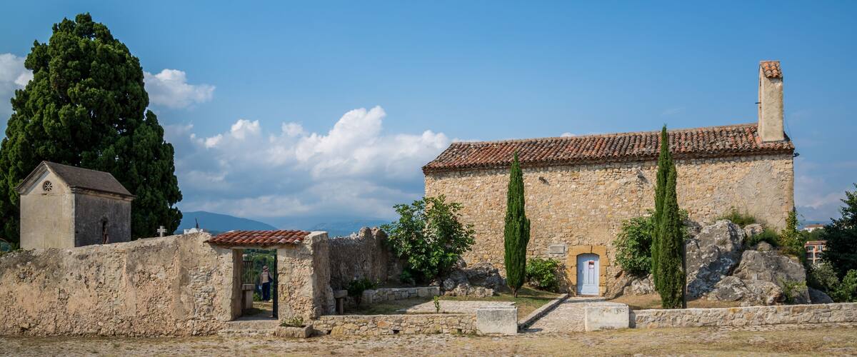 An ancient stone building with a tiled roof, a small church, and a large tree in a rural setting.