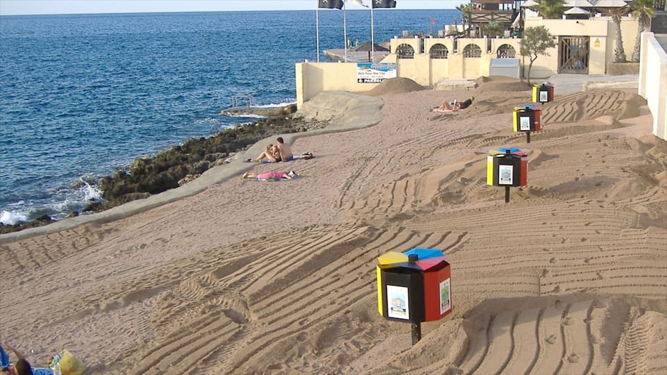 Une plage de sable avec des empreintes et une côte rocheuse. On aperçoit des poubelles colorées et un bâtiment orné d’arches au fond.