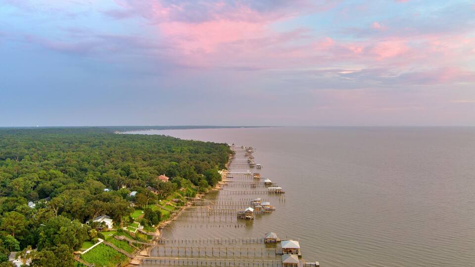 A long pier extending into a calm body of water, surrounded by lush greenery and a few small buildings.