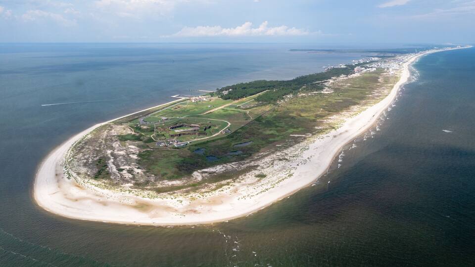 An aerial view of a coastal area with a long sandy beach, a small peninsula with buildings, and a clear view of the ocean.