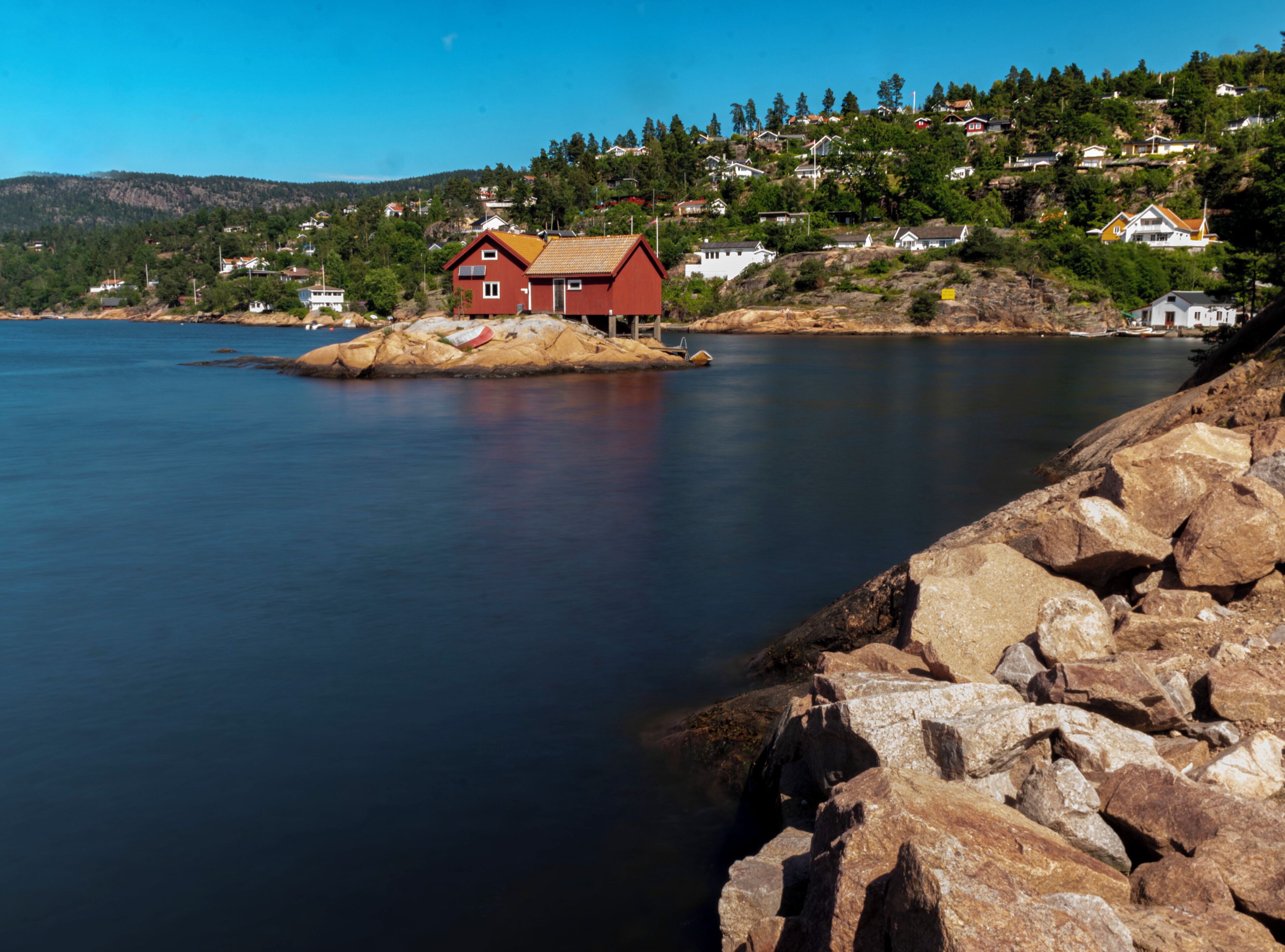 A red house on a small island in a calm body of water, surrounded by rocky shores and a hillside with houses.
