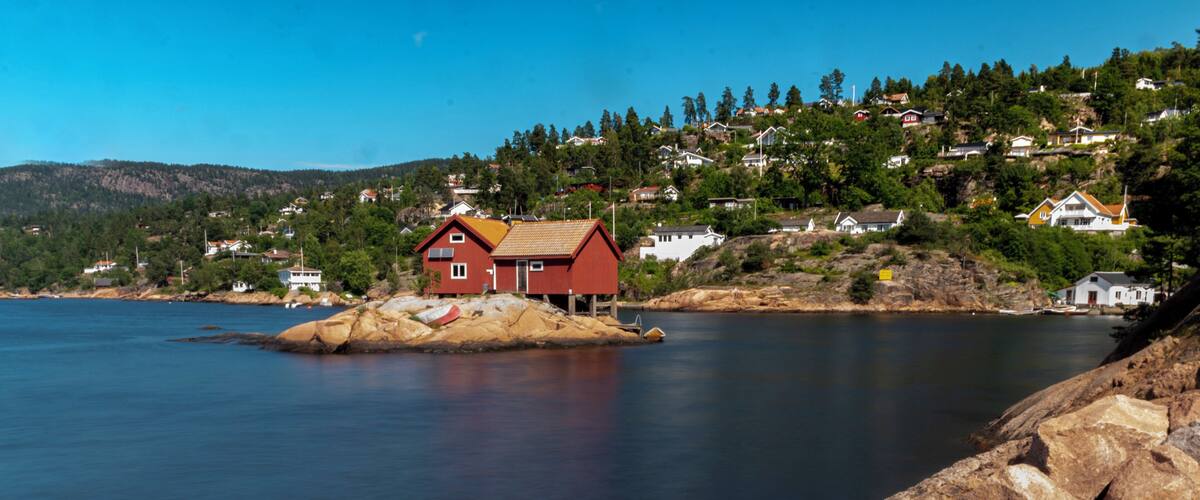 A red house on a small island in a calm body of water, surrounded by rocky shores and a hillside with houses.