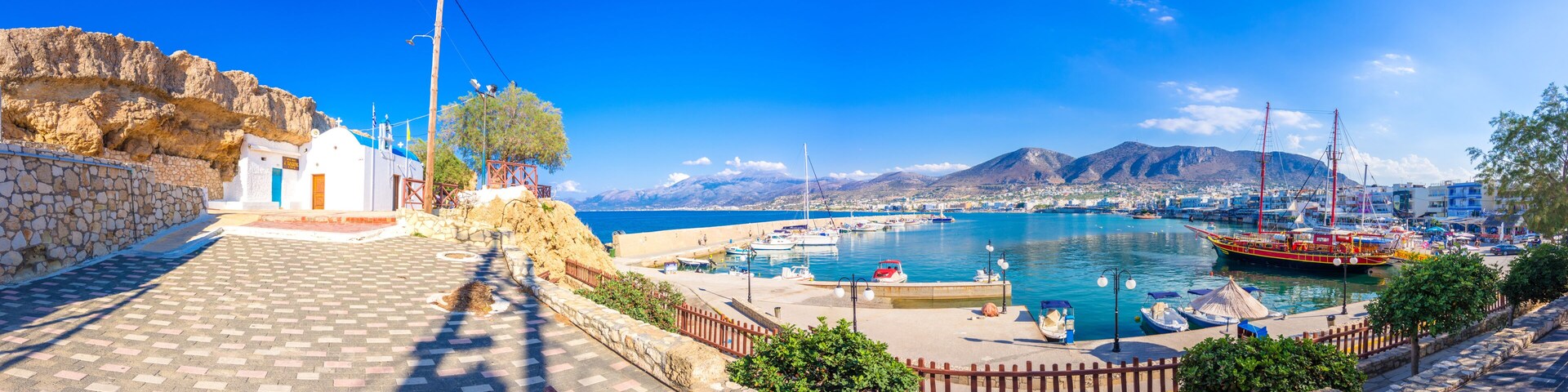 Un pueblo costero con un puerto, barcos, un muro de piedra y montañas al fondo.