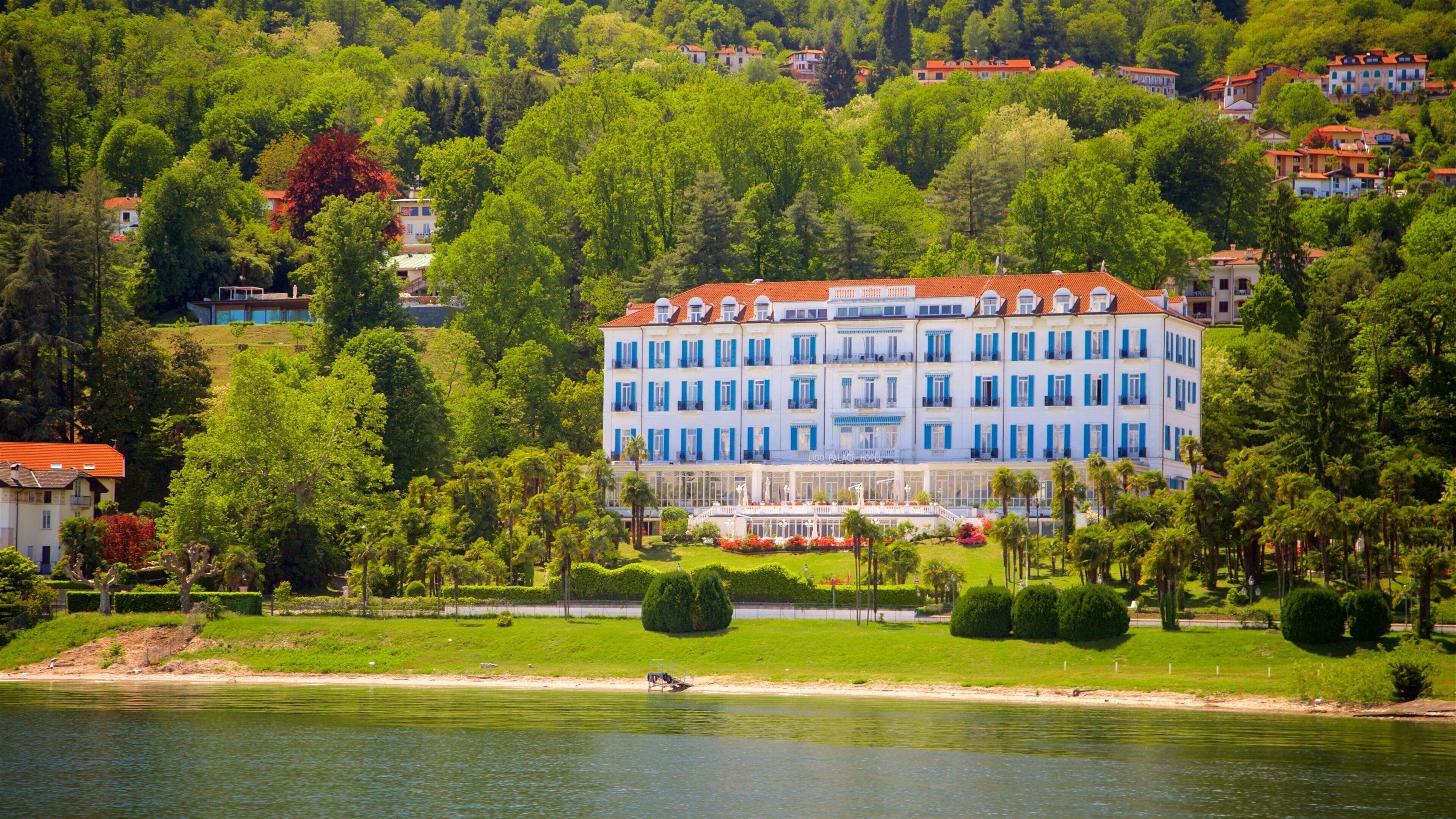 A large, multi-story hotel with blue shutters and a red roof, surrounded by lush greenery and a lake.