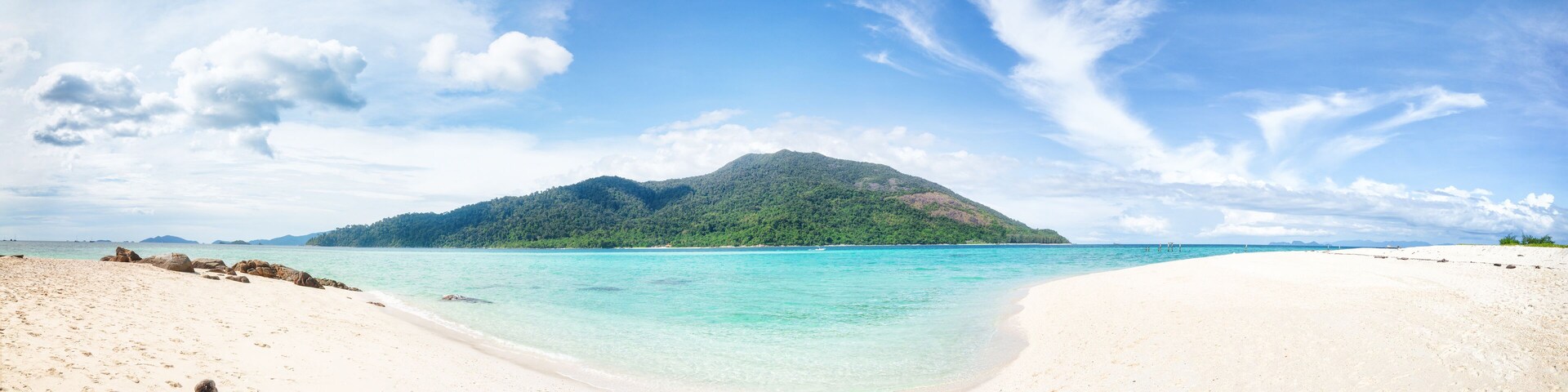 Ein Strand mit klarem türkisblauem Wasser, weißem Sand und einem grünen Berg im Hintergrund.