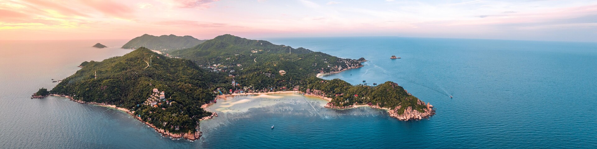 Vista aérea de una isla exuberante y verde con una playa, rodeada de aguas azules cristalinas y montañas distantes.