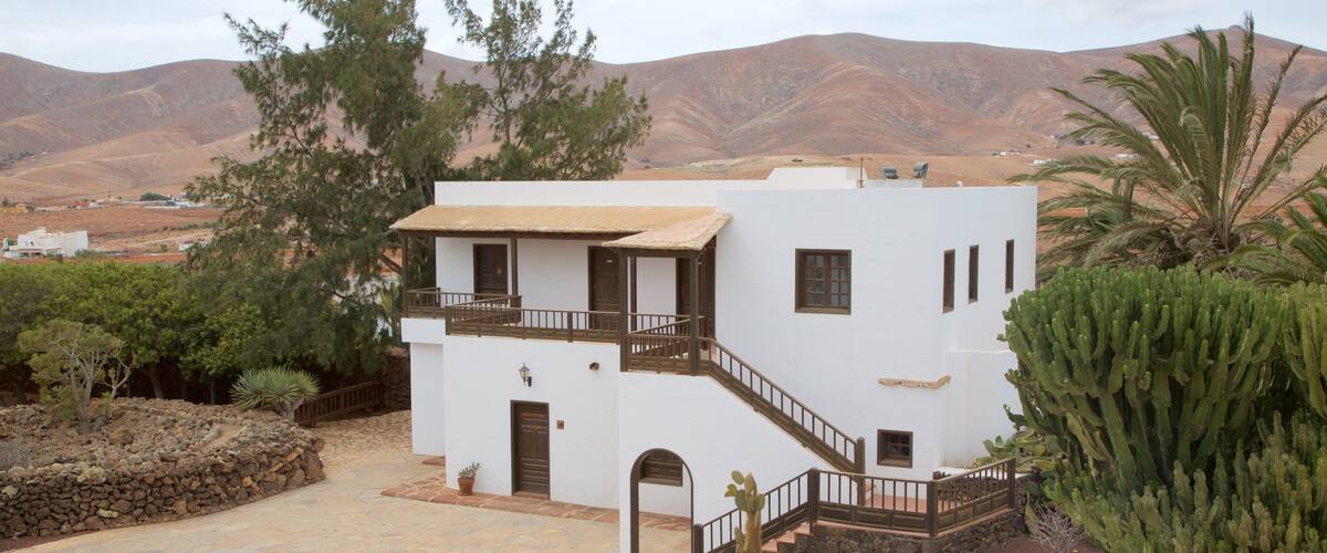 A white two-story house with a balcony, surrounded by cacti and desert vegetation.