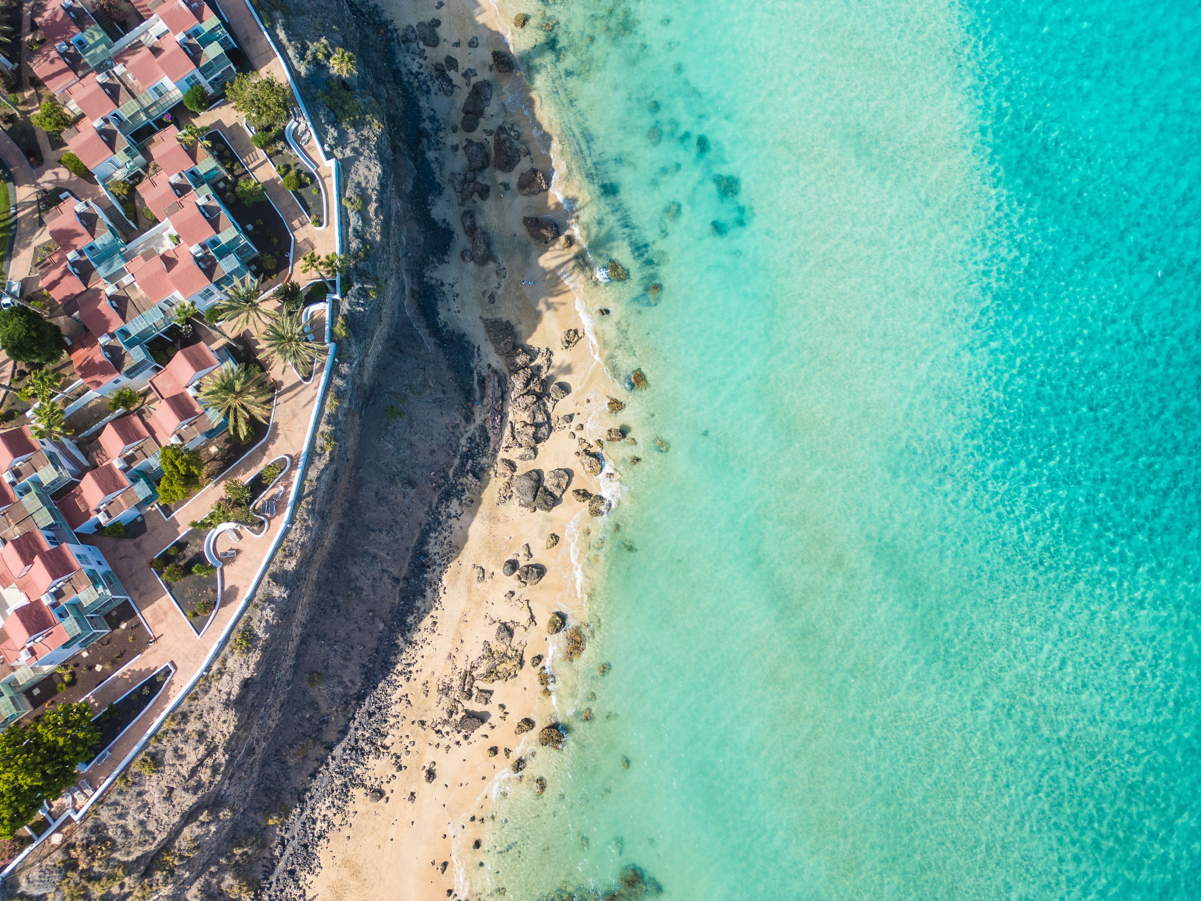 An aerial view of a coastal area with a sandy beach, clear turquoise waters, and a row of red-roofed buildings adjacent to the shore.