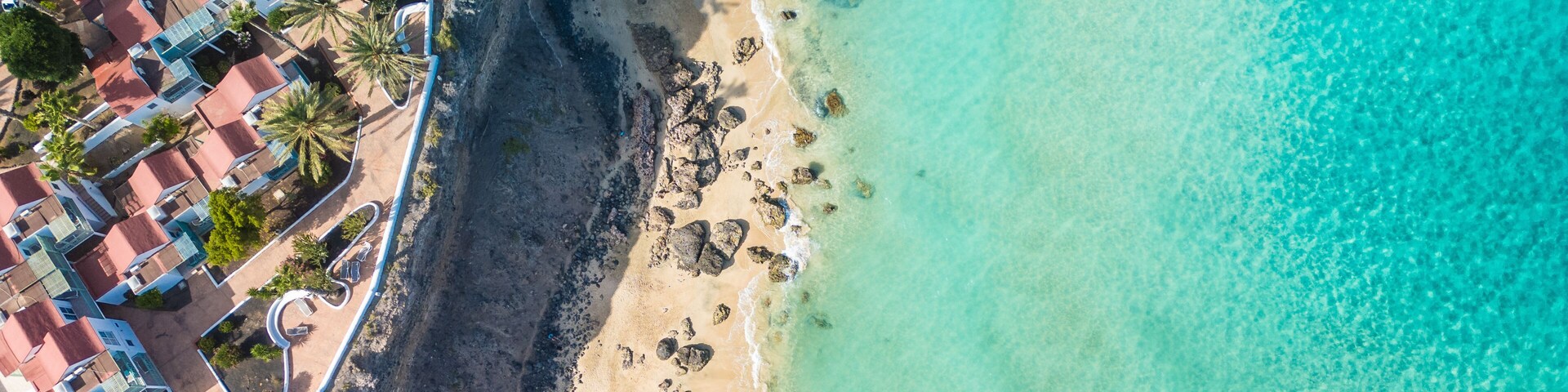 An aerial view of a coastal area with a sandy beach, clear turquoise waters, and a row of red-roofed buildings adjacent to the shore.