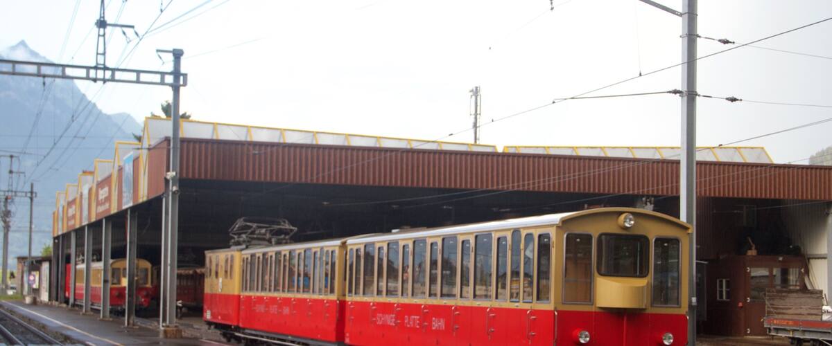 A red and yellow tram at a station with overhead wires and a mountainous backdrop.