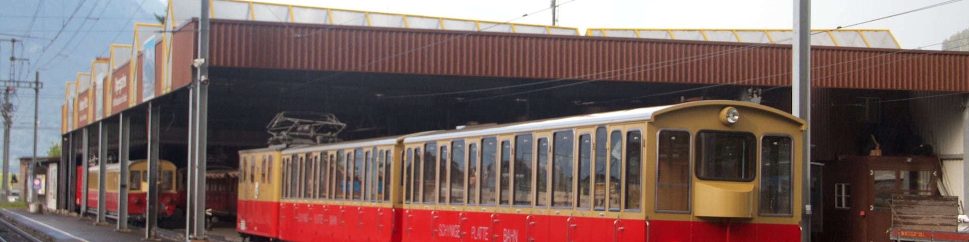 A red and yellow tram at a station with overhead wires and a mountainous backdrop.