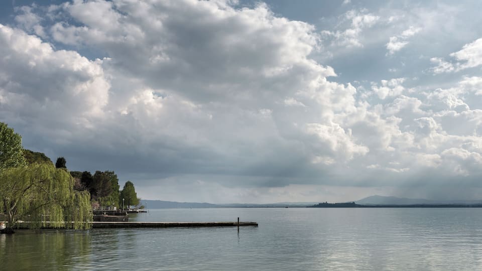 A calm lake with a dock, trees, and distant mountains under a cloudy sky.