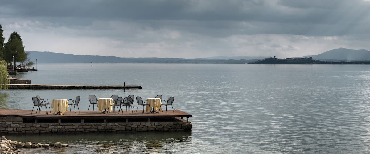 A lakeside deck with tables and chairs, a dock extending into the water, and distant mountains under a cloudy sky.