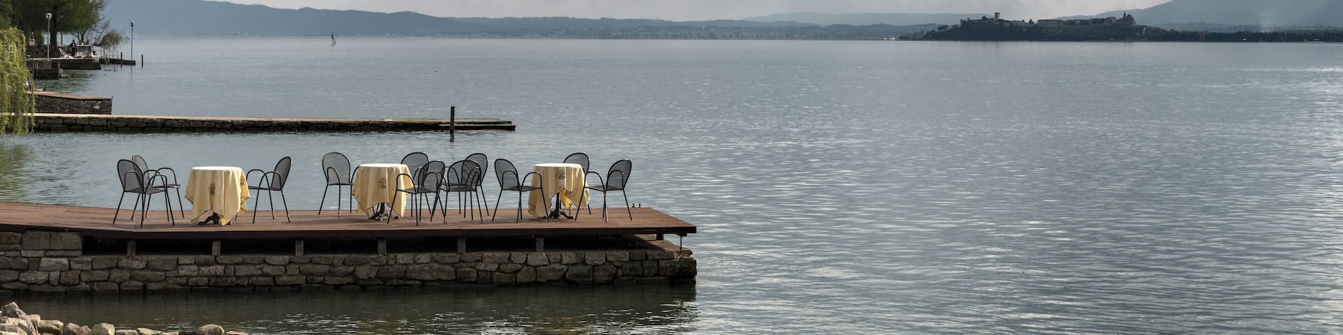 A lakeside deck with tables and chairs, a dock extending into the water, and distant mountains under a cloudy sky.
