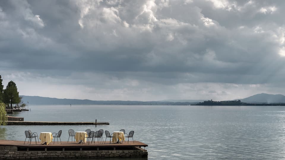 A lakeside deck with tables and chairs, a dock extending into the water, and distant mountains under a cloudy sky.