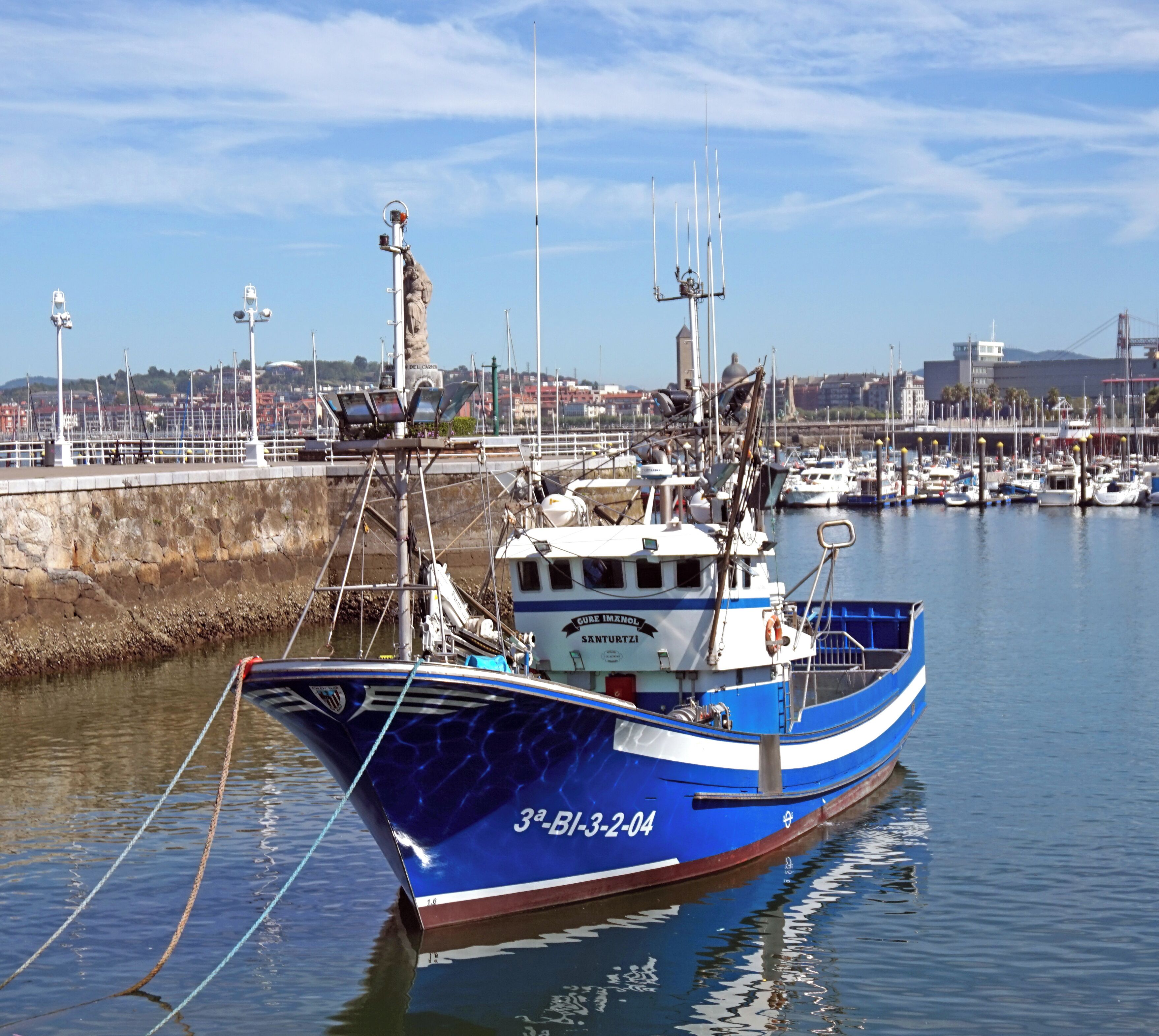 Un barco pesquero azul amarrado en un puerto deportivo, con un paisaje urbano al fondo.