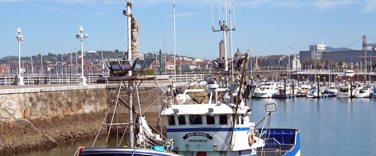 A blue fishing boat docked at a marina with a cityscape in the background.
