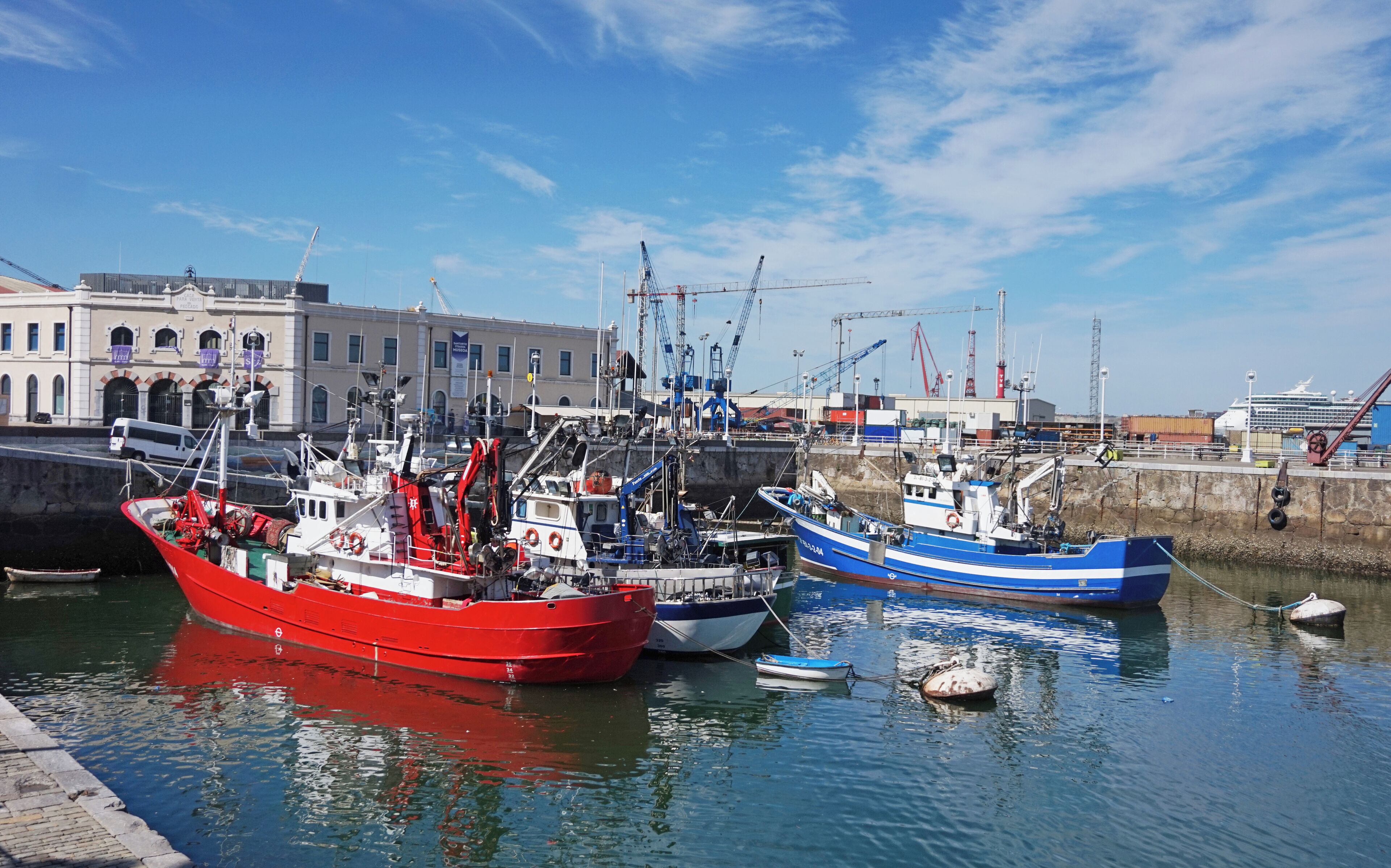 A harbor with several boats, including a red fishing boat and a blue boat, docked near a building with a sign that reads "Marina."