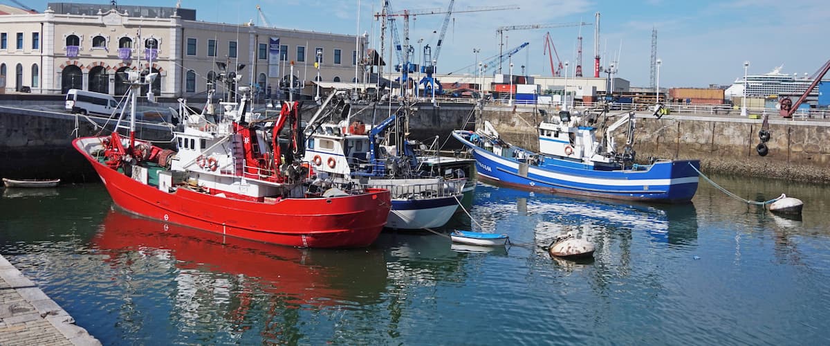 A harbor with several boats, including a red fishing boat and a blue boat, docked near a building with a sign that reads "Marina."